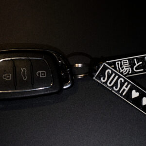 "A 3D-printed keychain attached to a black car key fob. The keychain consists of two rectangular tags: one featuring Japanese text and a star symbol, and the other displaying the word 'SUSH' with three heart symbols. The tags are black with white raised details, showcasing the precision of 3D printing."