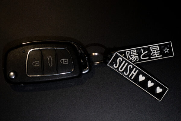 "A 3D-printed keychain attached to a black car key fob. The keychain consists of two rectangular tags: one featuring Japanese text and a star symbol, and the other displaying the word 'SUSH' with three heart symbols. The tags are black with white raised details, showcasing the precision of 3D printing."