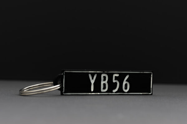 A rectangular 3D-printed keychain in black with raised white text reading 'YB56.' The keychain is attached to a metal keyring and placed against a dark background, showcasing the crisp details and precision of 3D printing.