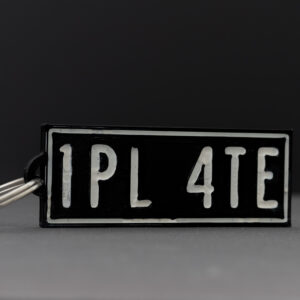 Close-up of a 3D printed black rectangular keychain with "1PL 4TE" in white text, photographed against a gray background.