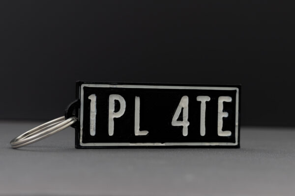 Close-up of a 3D printed black rectangular keychain with "1PL 4TE" in white text, photographed against a gray background.