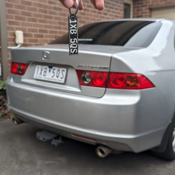 3d Printed Car key charm displaying "1XB 5QS" in black with white text, shown in front of a silver sedan with distinctive red taillights